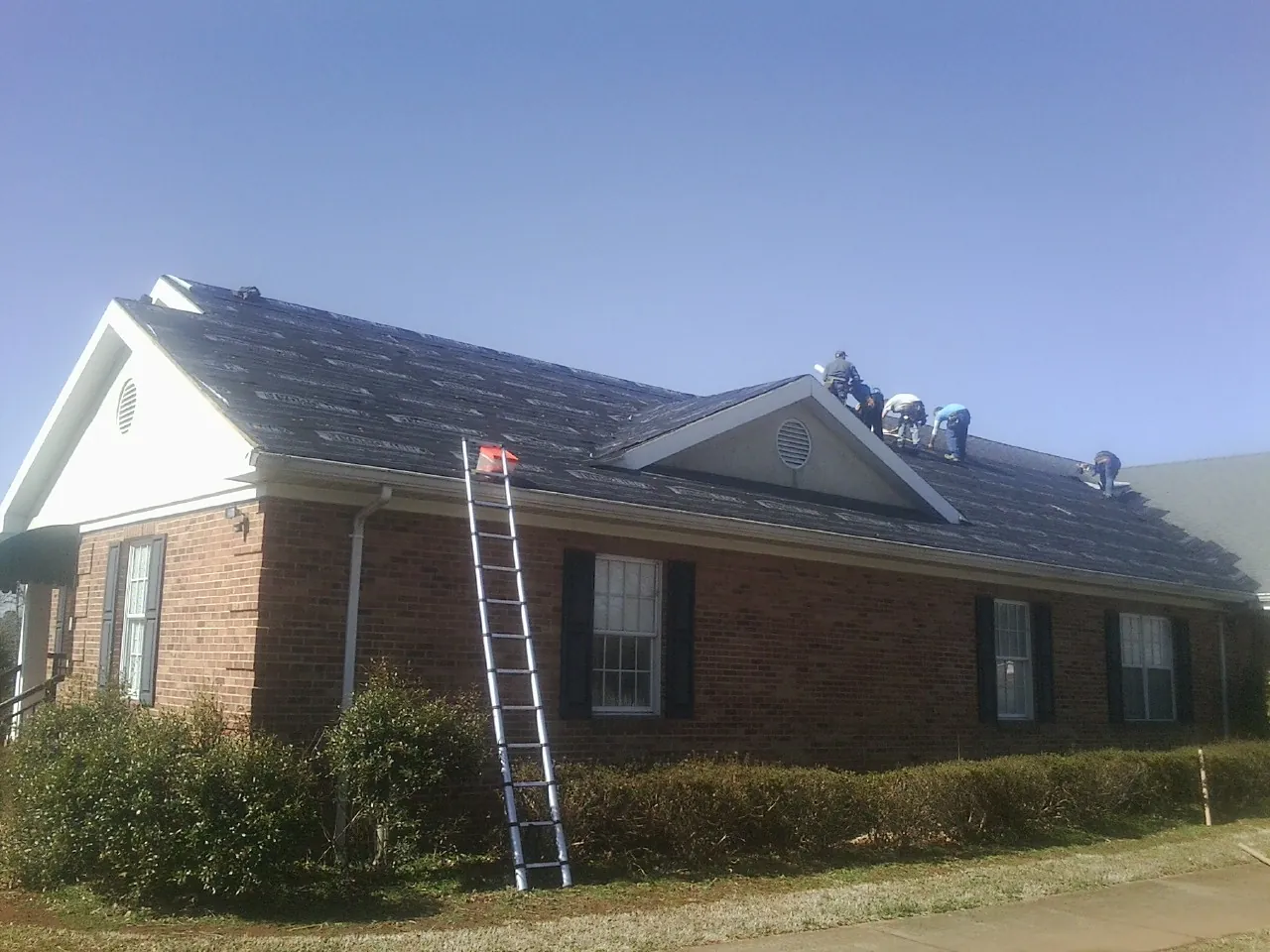 Crew tearing shingles off a brick ranch roof with a ladder set — Metal Roof Repair work by our crew in Waukee