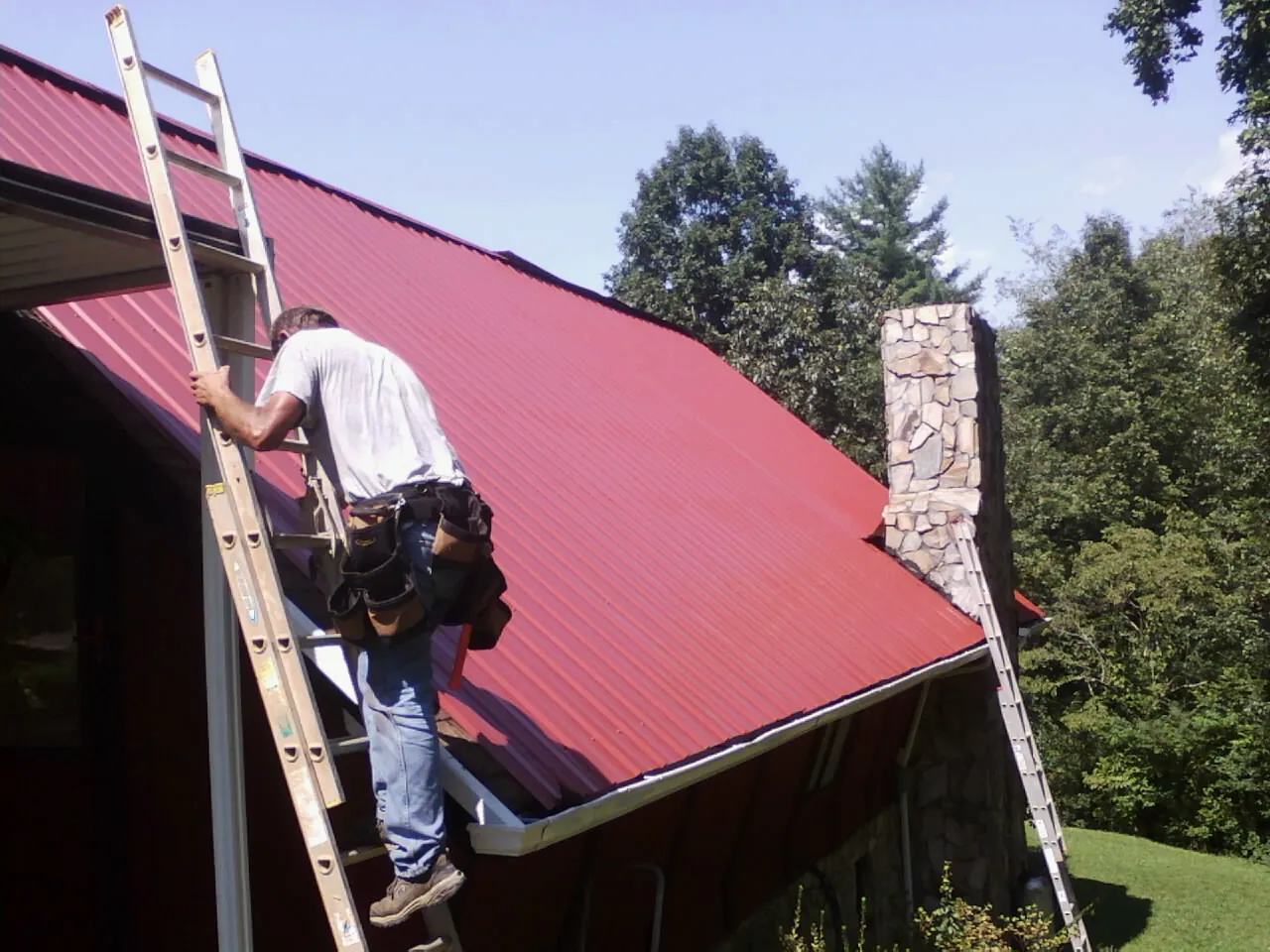 Roofer on a ladder beside a red metal roof and stone chimney — Soffit & Fascia Repair by Roofing Brothers in Waukee